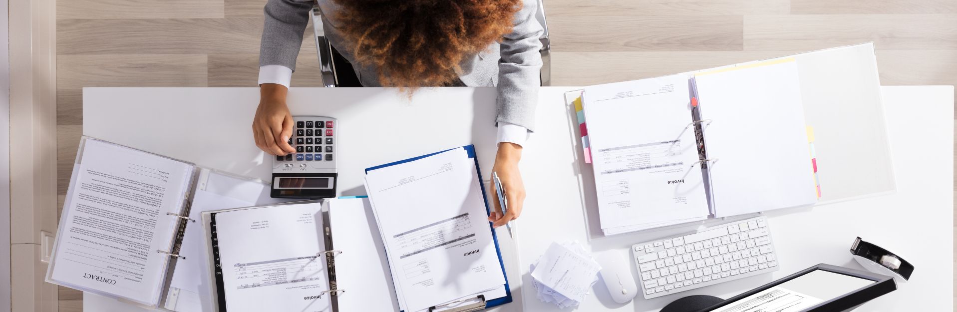 A nonprofit employee sits at a desk surrounded by invoices, contracts, and a calculator, reviewing financial documents and calculating figures.