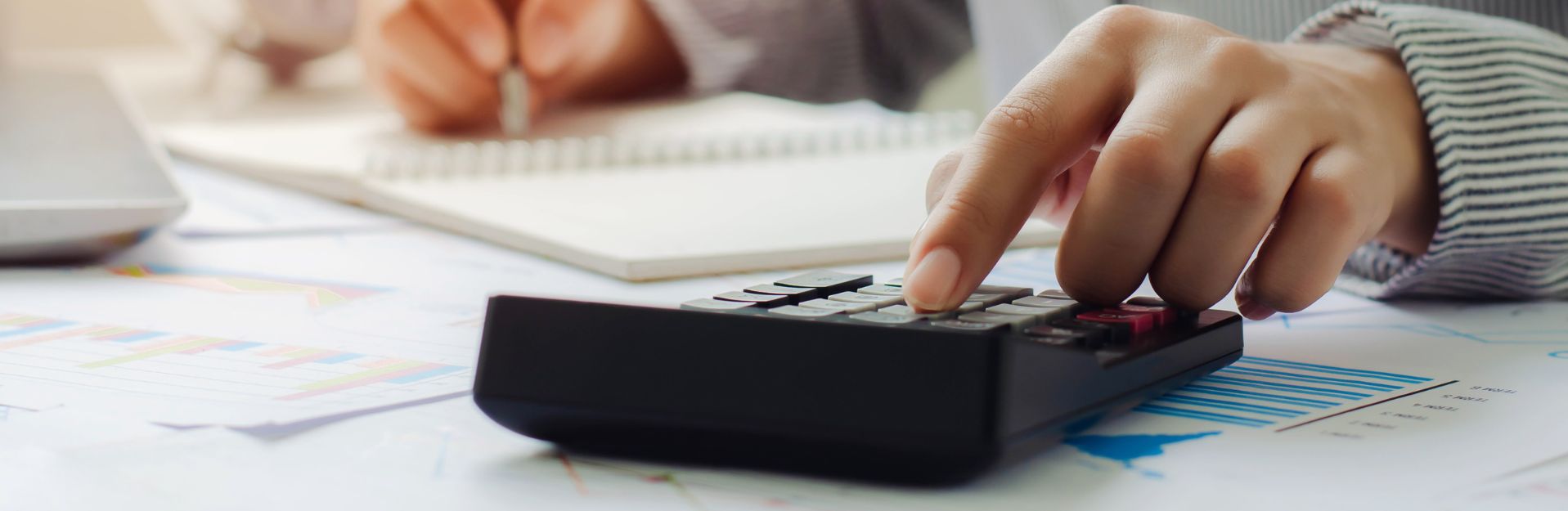 Close-up of a hand pressing a calculator, with colorful charts and a notebook in the background, indicating financial planning or budgeting.